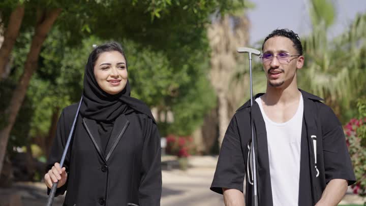 الاشارة الى شيء ما، الاستعداد للعب والتنافس، ممارسة الألعاب الحركية للترفيه في الهواء الطلق، قضاء وقت ممتع بالخارج، تصوير من الخلف لزوجان عربيان خليجيان يقومان بالتنزه فى الحدائق يمسكان بيدهم مضرب الجولف