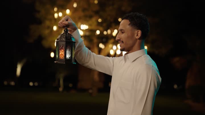 Gestures of happiness and joy, the nighttime Ramadan atmosphere, the joy of welcoming the month of Ramadan, a close-up shot of an Arab Gulf Saudi man wearing a white thobe holding a decorative metal lantern in his hands.