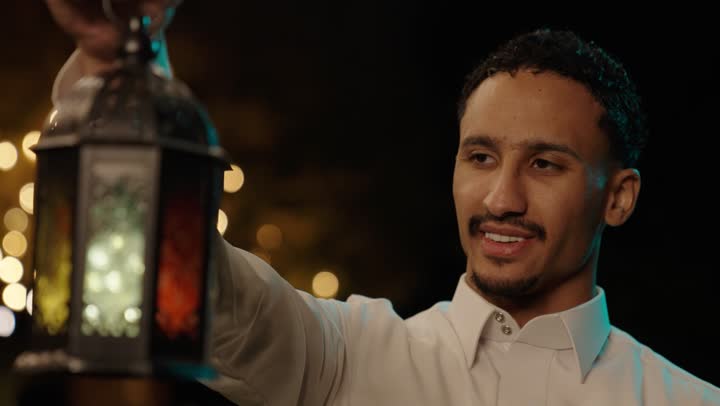 الأجواء الرمضانية الليلية، فرحة استقبال شهر رمضان، تصوير مقرب لرجل عربي خليجي سعودي يرتدي الثوب الابيض يمسك بيدية فانوس معدني مزخرف ، ايماءات السعادة و السرور