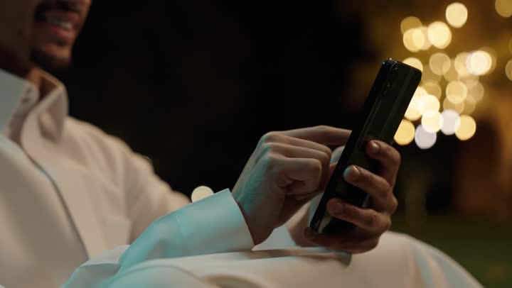 تصفح التطبيقات وبرامج التواصل الاجتماعي، استخدام جهاز تقني حديث ومتطور، العمل عن بعد، التواصل وتبادل الأحاديث والأخبار مع الأهل والأصدقاء، تصوير مقرب ليد رجل عربي خليجي سعودي يرتدي الثوب الابيض يجلس فى الهواء الطلق يمسك بيده الهاتف المحمول