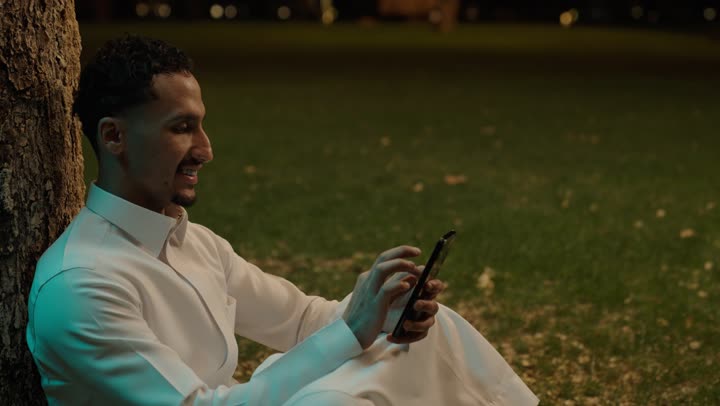استخدام جهاز تقني حديث ومتطور، العمل عن بعد، التواصل وتبادل الأحاديث والأخبار مع الأهل والأصدقاء، رجل عربي خليجي سعودي يرتدي الثوب الابيض يجلس فى الهواء الطلق يمسك بيده الهاتف المحمول، تصفح التطبيقات وبرامج التواصل الاجتماعي