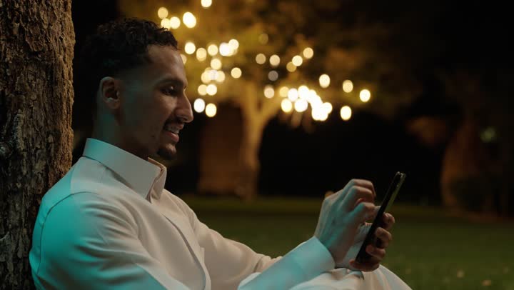 العمل عن بعد، التواصل وتبادل الأحاديث والأخبار مع الأهل والأصدقاء، رجل عربي خليجي سعودي يرتدي الثوب الابيض يجلس فى الهواء الطلق يمسك بيده الهاتف المحمول، استخدام جهاز تقني حديث ومتطور، تصفح التطبيقات وبرامج التواصل الاجتماعي