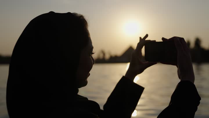 قضاء نزهه ترفيهية في النيل، تصوير مقرب من الخلف لامراه عربيه خليجيه سعودية محجبة ترتدي العباءة تمسك بيديها الهاتف المحمول، توثيق اللحظات السعيدة