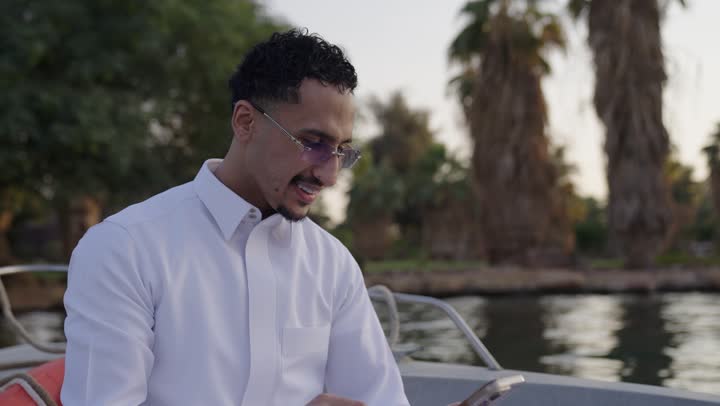 Communication and exchanging conversations and news with family and friends, a Gulf Arab Saudi man wearing a white thobe sitting inside a boat holding a mobile phone, using a modern and advanced technological device, working remotely, browsing applications and social media programs.