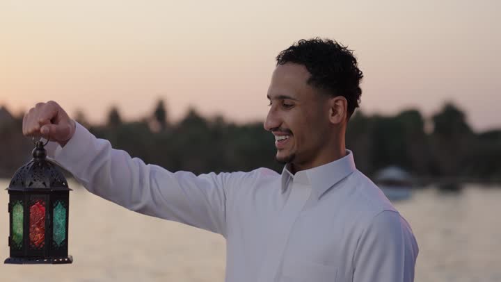 الاستمتاع بالأجواء الروحانية في لشهر المبارك، النظرإلى فانوس رمضان، إيماءات السعادة والسرور، العبادة والتقرب إلى الله، تصوير مقرب لرجل عربي سعودي خليجي يرتدي الثوب الابيض يقف فى الهواء الطلق يمسك بيدية الفانوس