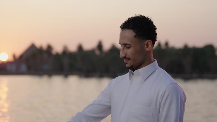 النظرإلى فانوس رمضان، إيماءات السعادة والسرور، العبادة والتقرب إلى الله، الاستمتاع بالأجواء الروحانية في لشهر المبارك، تصوير مقرب لرجل عربي سعودي خليجي يرتدي الثوب الابيض يقف فى الهواء الطلق يمسك بيدية الفانوس