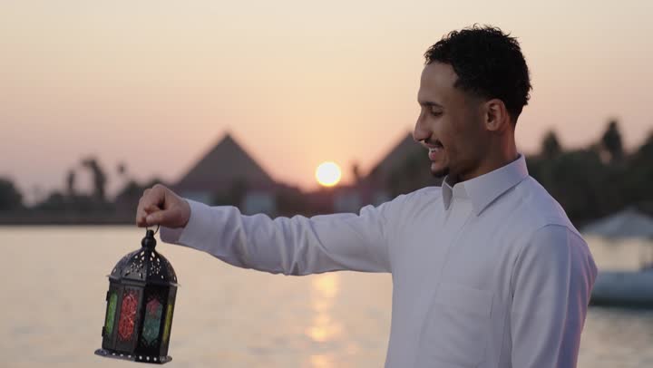 Gestures of happiness and joy, looking at the Ramadan lantern, worship and drawing closer to God, enjoying the spiritual atmosphere of the blessed month, a close-up shot of an Arab Saudi Gulf man wearing a white thobe standing outdoors holding the lantern in his hands.