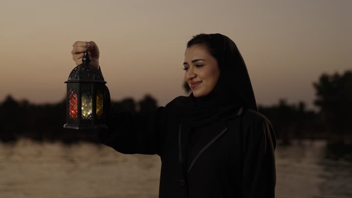 فوانيس وزينة الأعياد والمناسبات الاسلامية، العبادة والتقرب إلى الله، الإستعداد لقدوم شهر رمضان المبارك، أجواء السكينة والروحانية، امراة عربيه خليجيه سعودية محجبة ترتدي العباءة تقف فى الهواء الطلق تحمل بيدها فانوس معدني