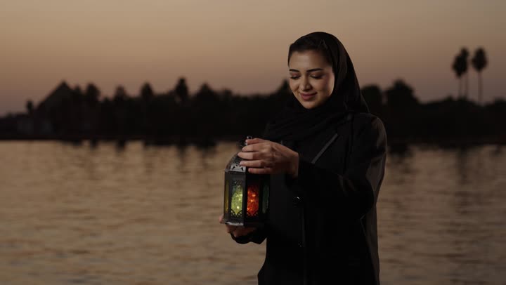 النظر للكاميرا بتعابير السعادة و السرور، العبادة والتقرب إلى الله، الإستعداد لقدوم شهر رمضان المبارك، المناسبات والأعياد الدينية، أجواء السكينة والروحانية، امراة عربيه خليجيه سعودية محجبة ترتدي العباءة تقف فى الهواء الطلق تحمل بيدها فانوس معدني