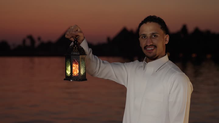 إضاءة وزينة شهر رمضان، تصوير مقرب لرجل عربي سعودي خليجي  يرتدي الثوب الابيض يقف فى الهواء الطلق يمسك فانوس شهر رمضان المبارك بايماءات وجه سعيدة، استقبال شهر رمضان المبارك