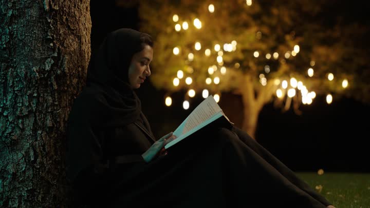 مطالعة القصص التصويرية المسلية، الجلوس على الأرض في إحدى الحدائق العامه، تمضية أوقات الفراغ بالتعلم والتثقف الذاتي، امراة عربيه خليجيه سعودية محجبة ترتدي العباءة تقوم بقراءة كتاب في الهواء الطلق