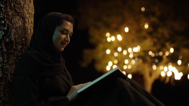 الجلوس على الأرض في إحدى الحدائق العامه، تمضية أوقات الفراغ بالتعلم والتثقف الذاتي، امراة عربيه خليجيه سعودية محجبة ترتدي العباءة تقوم بقراءة كتاب في الهواء الطلق
