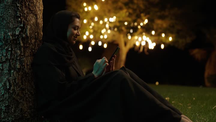 تصفح التطبيقات وبرامج التواصل الاجتماعي، استخدام جهاز تقني حديث ومتطور، امراة عربية خليجية سعودية مبتسمة ترتدي حجاب وعباءة تجلس فى الهواء الطلق تمسك بيديها الهاتف المحمول، الدردشة والتواصل مع التواصل مع الأصدقاء اونلاين