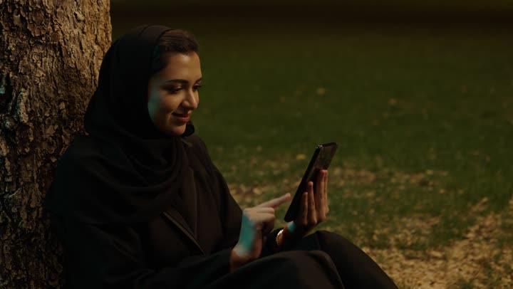 Using a modern and advanced technical device, browsing social media using a mobile phone, a smiling Arab Gulf Saudi woman wearing a hijab and abaya sitting outdoors holding a mobile phone in her hands, chatting and communicating with friends online.