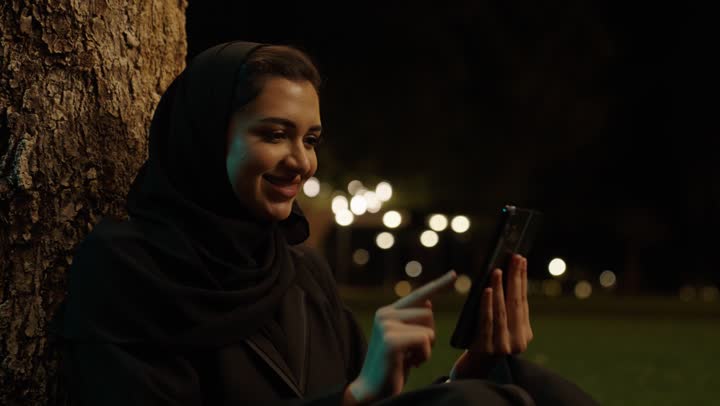 الدردشة والتواصل مع التواصل مع الأصدقاء اونلاين، تعابير وايماءات الفرح والسرور، تصفح وسائل التواصل الاجتماعي باستخدام الجوال،تصوير مقرب لإمرأة عربية خليجية سعودية مبتسمة ترتدي حجاب وعباءة  تمسك بيديها الهاتف المحمول