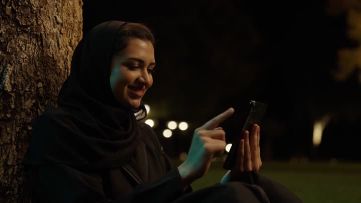 تعابير وايماءات الفرح والسرور، تصفح وسائل التواصل الاجتماعي باستخدام الجوال،تصوير مقرب لإمرأة عربية خليجية سعودية مبتسمة ترتدي حجاب وعباءة  تمسك بيديها الهاتف المحمول، الدردشة والتواصل مع التواصل مع الأصدقاء اونلاين