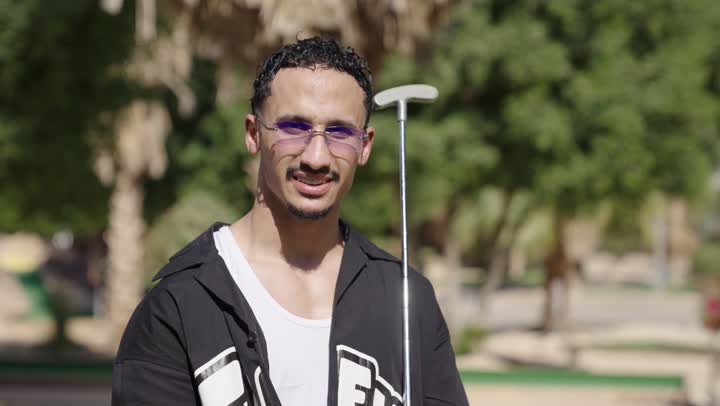 اللعب الحركي والاستمتاع في الهواء الطلق، رجل عربي سعودي خليجي يرتدي الزي الكاجوال يمسك مضرب الجولف، الاستعداد للعب والتنافس، النظر للكاميرا بايماءات السعادة و السرور