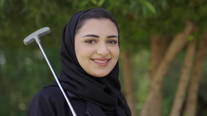 الترفيه بالأنشطة الحركية الخارجية، الاستعداد للعب والتنافس، النظر للكاميرا بتعابير السرور، قضاء وقت ممتع بالخارج، تصوير مقرب لامراة عربيه خليجيه سعودية محجبة ترتدي العباءة تمسك بيديها مضرب الجولف