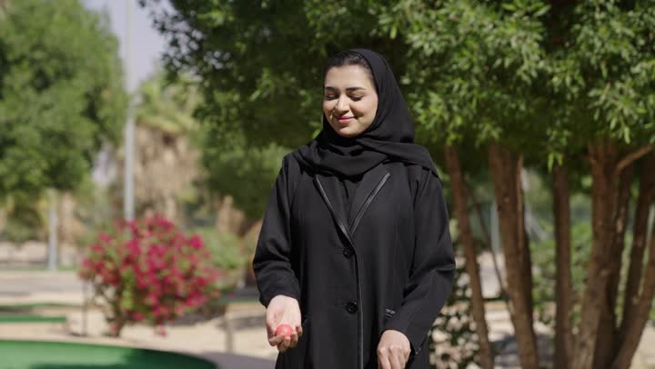 Looking at the camera with expressions of joy, having fun and entertainment with outdoor physical games, spending enjoyable time outside, an Arab Gulf Saudi woman wearing a hijab and an abaya hitting a golf ball several times and holding the club, getting ready to play and compete.