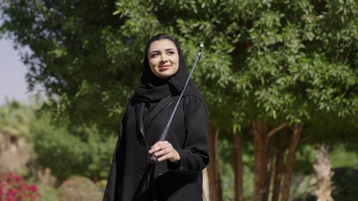 قضاء وقت ممتع بالخارج، امرأة عربية خليجية سعودية محجبة ترتدي العباءة تتجول في الحديقة الخضراء وتمسك مضرب جولف، النظر الى شيء ما، تعابير السعادة و السرور