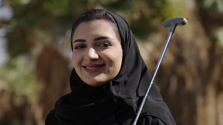 Enjoying a good time outdoors, playing golf, a close-up shot of a young Arab Gulf Saudi woman wearing a hijab and an abaya holding a golf club, summer Olympic sports, looking at the camera with expressions of happiness and joy.