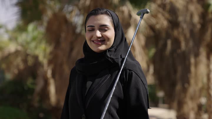 النظر للكاميرا بتعابير السعادة و السرور، الاستمتاع بقضاء وقت ممتع فى الخارج، ممارسة لعبة الغولف، تصوير مقرب  لشابة عربيه خليجيه سعودية محجبة ترتدي العباءة تمسك بيديها مضرب الغولف، رياضات الألعاب الأولمبية الصيفية