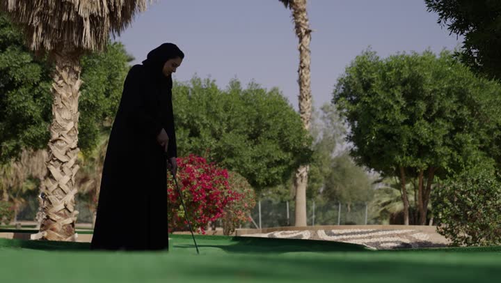 الاستمتاع بلعبة الجولف في الملعب، ممارسة لعبة الغولف في مساحات واسعة خضراء، شابة عربيه خليجيه سعودية محجبة ترتدي العباءة تمسك المضرب و تقوم بضرب الكرة بعيدا، نشاط رياضي بالمملكة العربية السعودية، رياضات الألعاب الأولمبية الصيفية
