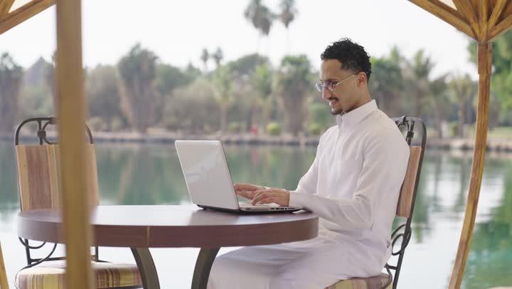 صفح الانترنت باستخدام الابتوب، الكتابة على لوحة المفاتيح، العمل عن بعد، شاب عربي خليجي سعودي يرتدي الثوب الابيض يجلس في المقهى الخارجي ويستخدم جهاز الحاسوب المحمول بايماءات السرور