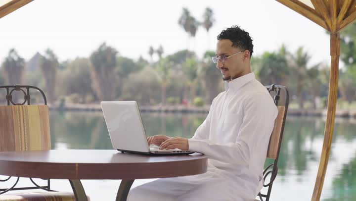 الاستمتاع بالاجواء الهادئه خارج المنزل، الكتابة على لوحة المفاتيح، العمل عن بعد، شاب عربي خليجي سعودي يرتدي الثوب الابيض يجلس في المقهى الخارجي ويستخدم جهاز الحاسوب المحمول بايماءات السرور