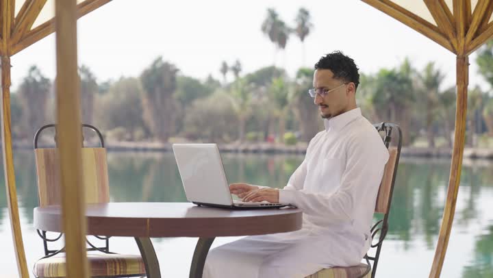 Remote work, using modern and advanced technical devices to complete projects in the cafe, typing on the keyboard, a young Arab Gulf Saudi man wearing a white thobe sitting in an outdoor cafe and using a laptop with gestures of joy.