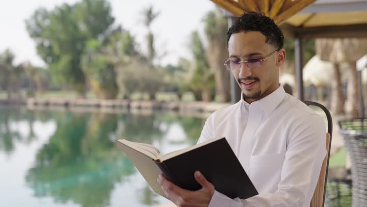 ثقافة التعلم الذاتي، حب المطالعة والقراءة ، قضاء أوقات هادئة في الخارج، ايماءات التركيز والاستمتاع، قراءة الروايات والقصص، تصوير مقرب لشاب عربي خليجي سعودي يرتدي الثوب التقليدي يحمل بيده كتاب