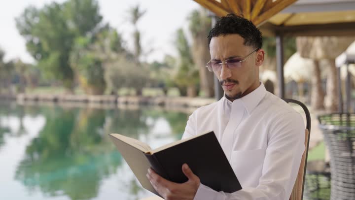 حب المطالعة والقراءة ، ثقافة التعلم الذاتي، قضاء أوقات هادئة في الخارج، ايماءات التركيز والاستمتاع، قراءة الروايات والقصص، تصوير مقرب لشاب عربي خليجي سعودي مبتسم يرتدي الثوب التقليدي يحمل بيده كتاب