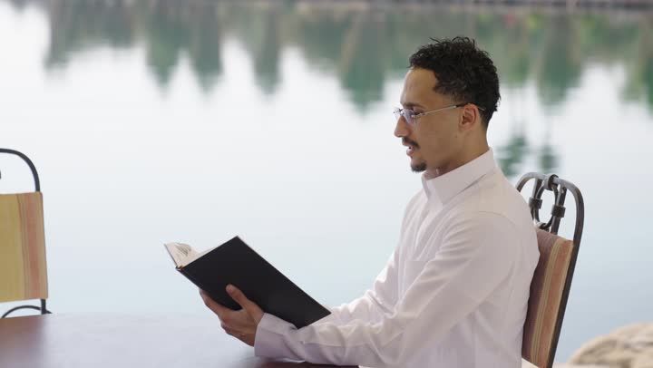 تمضية أوقات الفراغ بالتعلم والتثقيف الذاتي، قضاء أوقات هادئة في الخارج، ايماءات التركيز والاستمتاع، ممارسة هواية المطالعة، قراءة الروايات والقصص، تصوير مقرب لشاب عربي خليجي سعودي مبتسم يرتدي الثوب التقليدي يحمل بيده كتاب