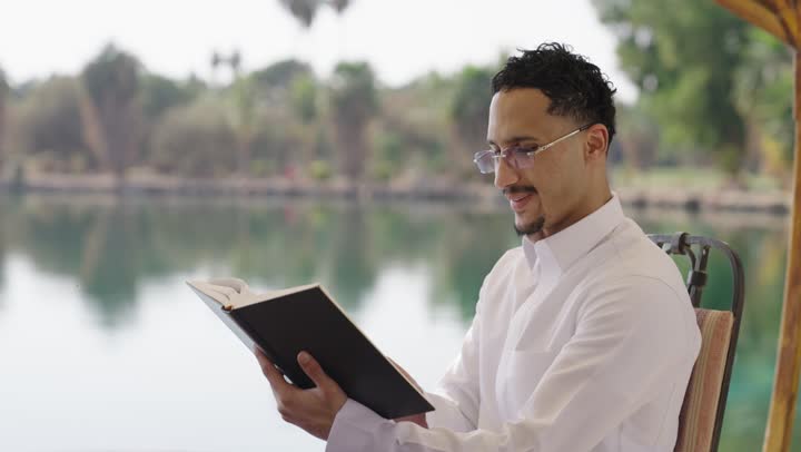 ايماءات التركيز والاستمتاع، ممارسة هواية المطالعة، قراءة الروايات والقصص، قضاء أوقات هادئة في الخارج، تمضية أوقات الفراغ بالتعلم والتثقيف الذاتي، تصوير مقرب لشاب عربي خليجي سعودي مبتسم يرتدي الثوب التقليدي يحمل بيده كتاب