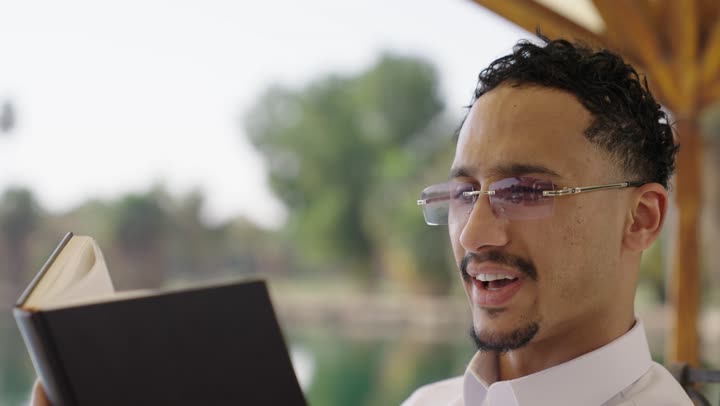 Practicing the hobby of reading, reading novels and stories, gestures of focus and enjoyment, spending quiet times outdoors, spending free time learning and self-education, a close-up shot of a smiling young Arab Gulf Saudi man wearing traditional attire holding a book in his hand.
