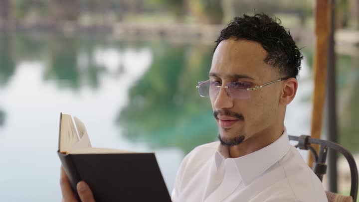 قراءة الروايات والقصص، ممارسة هواية المطالعة، ايماءات التركيز والاستمتاع، قضاء أوقات هادئة في الخارج، تمضية أوقات الفراغ بالتعلم والتثقيف الذاتي، تصوير مقرب لشاب عربي خليجي سعودي يرتدي الثوب التقليدي يحمل بيده كتاب