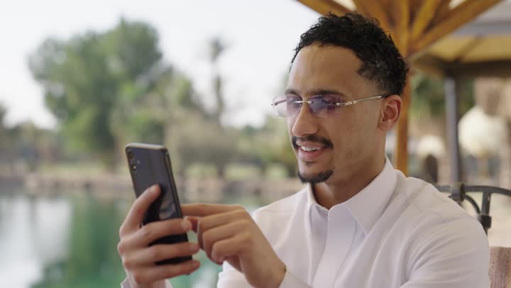 استخدام الأجهزة التقنية الحديثة للتسلية و الترفيه,تصفح تطبيقات الجوال، الدردشة والتحدث مع الاصدقاء عبر الانترنت, تصوير مقرب لشاب عربي خليجي سعودي يرتدي الثوب الابيض يجلس في احد المقاهي الخارجية، يستخدم الهاتف المحمول