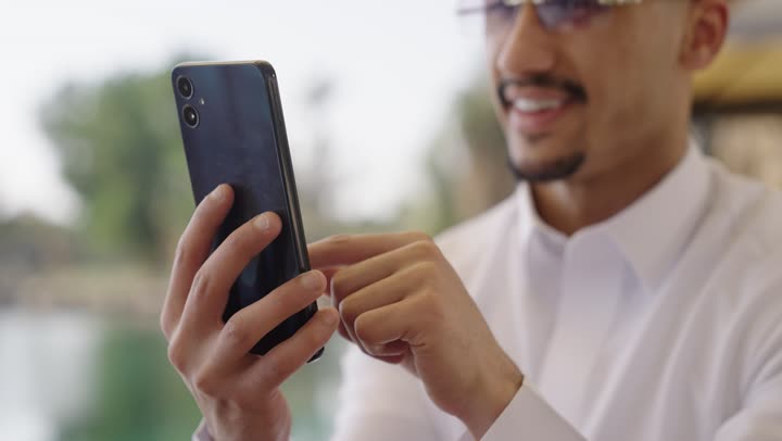 تصفح تطبيقات الجوال، دمج التقنيات الحديثة بالحياة اليومية، الدردشة والتحدث مع الاصدقاء عبر الانترنت, تصوير مقرب لشاب عربي خليجي سعودي يرتدي الثوب الابيض يجلس في احد المقاهي الخارجية، يستخدم الهاتف المحمول
