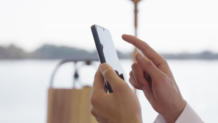 الدردشة والتحدث مع الاصدقاء عبر الانترنت، دمج التقنيات الحديثة بالحياة اليومية، التصفح عبر وسائل التواصل الاجتماعي, تصوير مقرب ليد شاب عربي خليجي سعودي يرتدي الثوب الابيض يجلس في احد المقاهي الخارجية، يستخدم الهاتف المحمول