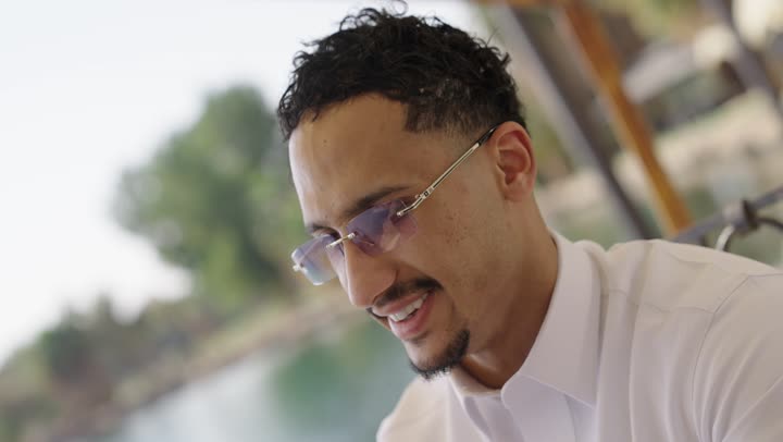 استخدام التكنولوجيا و التقنيات الحديثة، دمج التقنيات الحديثة بالحياة اليومية، التصفح عبر وسائل التواصل الاجتماعي, تصوير مقرب لشاب عربي خليجي سعودي يرتدي الثوب الابيض يجلس في احد المقاهي الخارجية، يستخدم الهاتف المحمول