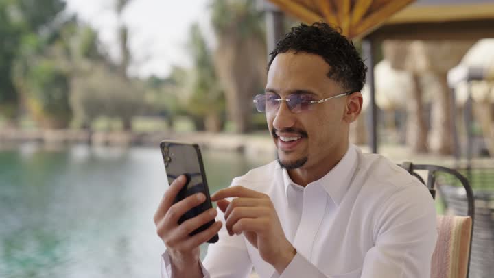 Integrating modern technologies into daily life, browsing through social media, using technology and modern techniques, a close-up shot of a young Arab Gulf Saudi man wearing a white thobe sitting in an outdoor cafe, using a mobile phone.