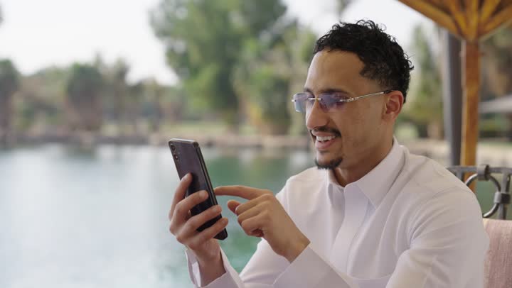 تصفح وسائل التواصل الاجتماعي، دمج التقنيات الحديثة في الحياة اليومية، استخدام التكنولوجيا والتقنيات الحديثة، لقطة قريبة لرجل سعودي شاب من الخليج العربي يرتدي ثوبًا أبيض ويجلس في مقهى خارجي يستخدم هاتفًا محمولًا.