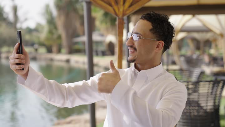 Communicating with friends and relatives through modern and advanced applications and technologies, making a video call with gestures of joy over the internet, a portrait of a smiling Saudi Arabian Gulf man wearing traditional attire sitting in an outdoor cafe using a mobile phone.