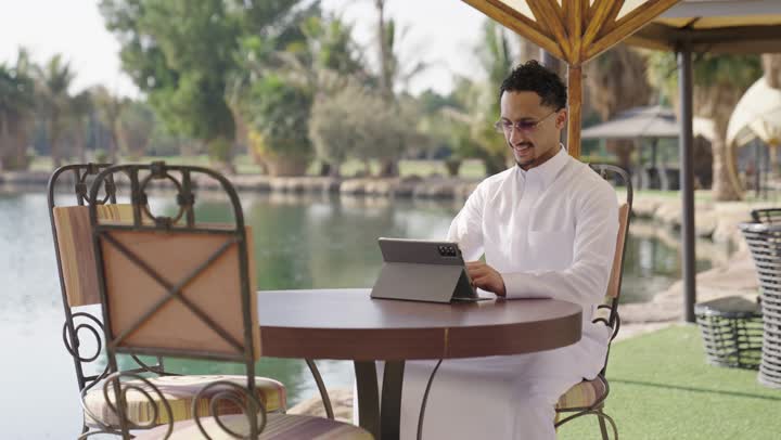 متابعة أخبار مواقع التواصل الاجتماعي، مفهوم التكنولوجيا وإستخدام تقنيات حديثة، استخدم جهاز التابلت لانجاز المشاريع والمهام، شاب عربي خليجي  سعودي يرتدي الثوب الابيض يجلس في احد المقاهي الخارجية، قضاء وقت ممتع فى الخارج