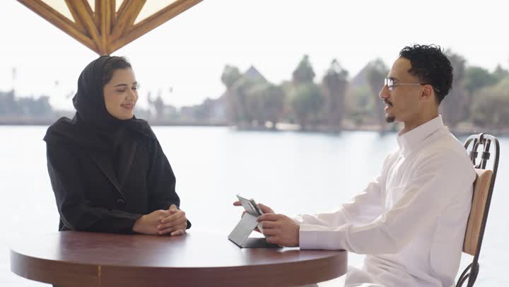 مطاعم وكافيهات راقية في السعودية، اجواء عائلية سعيدة، استخدام جهاز تقني حديث، زوجان عربيان خليجيان سعوديان يرتديان الزي التقليدي  يجلسان فى احد المقاهي الخارجية يقومان بمشاهده شيء ما على جهاز التابلت، نقاشات و حوارات زوجية