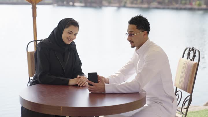 توثيق اللحظات وبناء علاقات اجتماعية ناجحة، التقاط صور السيلفي التذكارية باستخدام الهاتف المحمول، تقوية العلاقات الزوجية وروابط الحب والمودة، زوجان عربيان خليجيان اماراتيان يرتديان الزي التقليدي يجلسان فى احد المقاهي الخارجية، تعابير السعادة و السرور