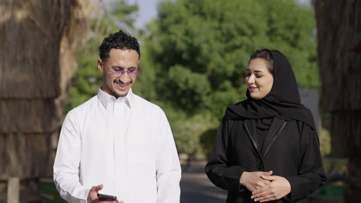 Family recreational activities, chatting and marital conversations, a Saudi Arabian Gulf couple dressed in traditional attire taking a selfie with a mobile phone, spending a summer vacation, an outdoor picnic, gestures of happiness and joy.