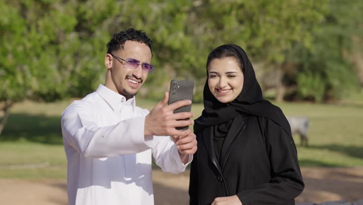 قضاء عطلة صيفية ،الاشارة الى شي ما، زوجان عربيان خليجيان سعوديان يرتدي الزي التقليدي يقومان بالتقاط صورة سيلفي عبر الهاتف المحمول، نزهه في الهواء الطلق، ايماءات السعادة و السرور، أنشطة ترفيهية عائلية