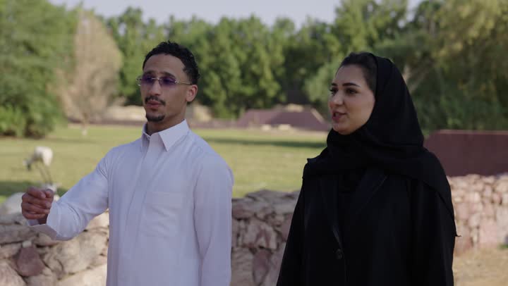 اجواء عائلية سعيدة، الاشارة الى شيء ما،زوجان عربيان خليجيان سعوديان يرتدي الزي التقليدي يقضيان أوقات ممتعة في إحدى الحدائق في السعودية، دردشة ومحادثات زوجية، قضاء عطلة صيفية، نزهه في الهواء الطلق، المشي في الطبيعة الخضراء، أنشطة ترفيهية عائلية