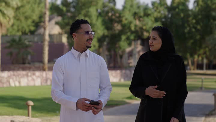 التنزه في الهواء الطلق، اجواء مليئة بالمحبة والود، مفهوم العلاقات الزوجية الناجحة السعيده، قضاء عطلة نهاية الاسبوع في أحد الحدائق ، رجل عربي خليجي سعودي يرتدي الثوب الابيض يقوم بتصفح الهاتف المحمول برفقة زوجته